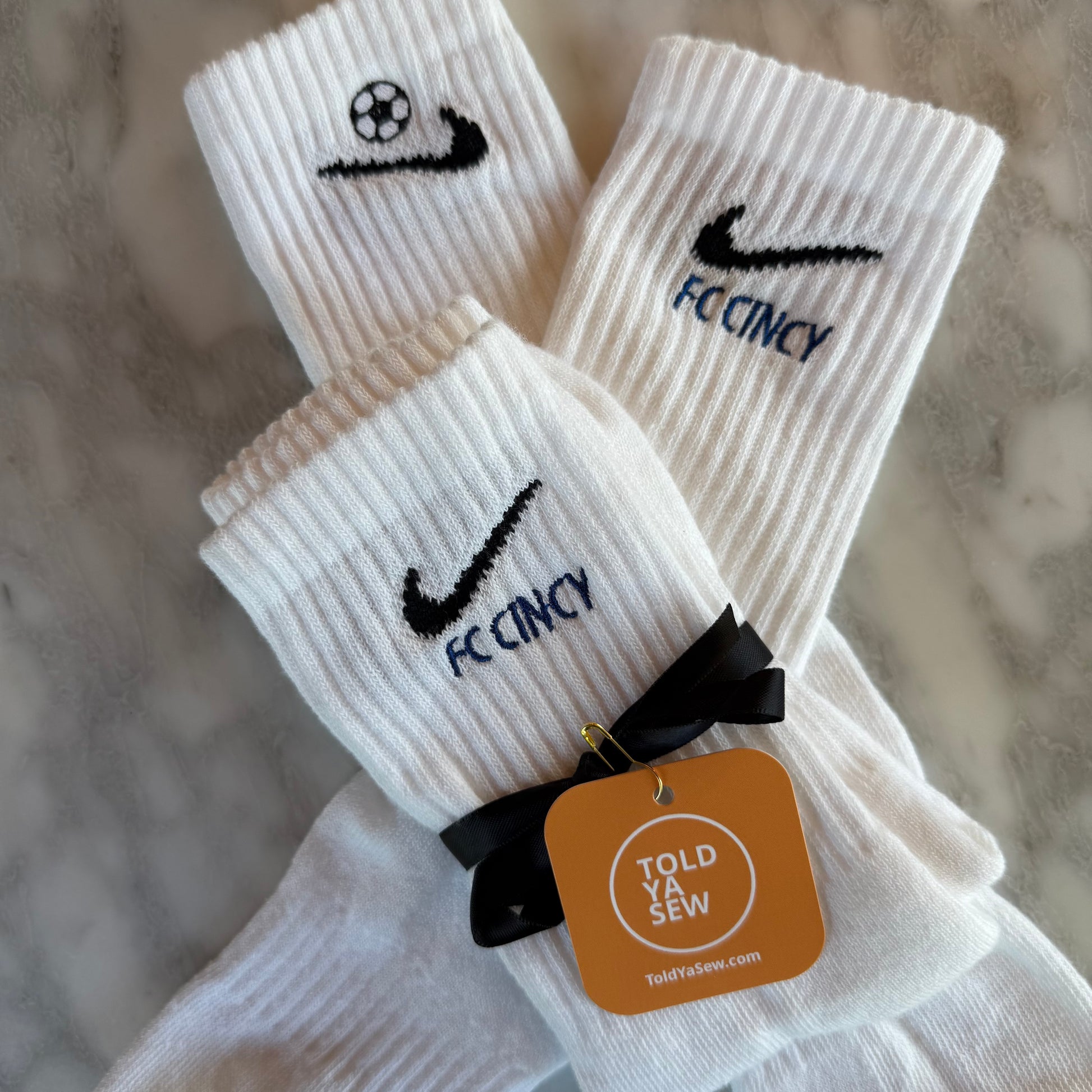 A pair of white Nike crew socks with 'FC Cincy' and a soccer ball embroidered on them.